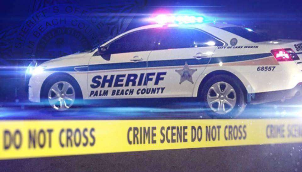 A Palm Beach County Sheriff's vehicle with flashing lights is parked at a crime scene, cordoned off with yellow tape that reads "CRIME SCENE DO NOT CROSS." The scene is illuminated by blue lights, indicating an active investigation.