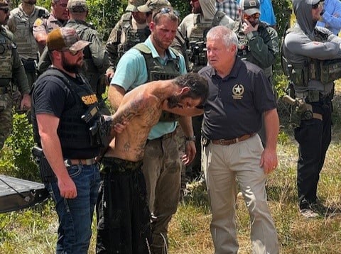 A shirtless man with tattoos is being escorted by law enforcement officers in a wooded area. The man is handcuffed, and a sheriff in a uniform stands nearby. Several other officers in tactical gear are present in the background, observing the scene.