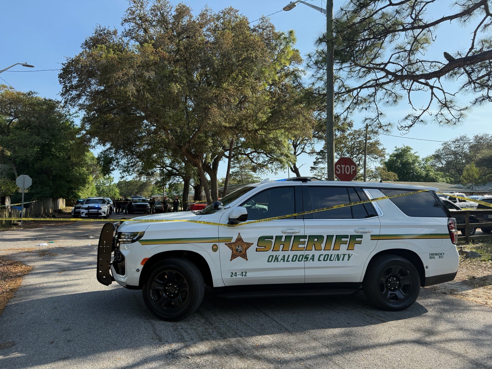 An Okaloosa County Sheriff's Office SUV is parked on a street near Shirley Drive and Marler Street in Fort Walton Beach, with yellow crime scene tape visible around the area. Multiple sheriff's vehicles and personnel are in the background, along with trees, a stop sign, and a clear blue sky.