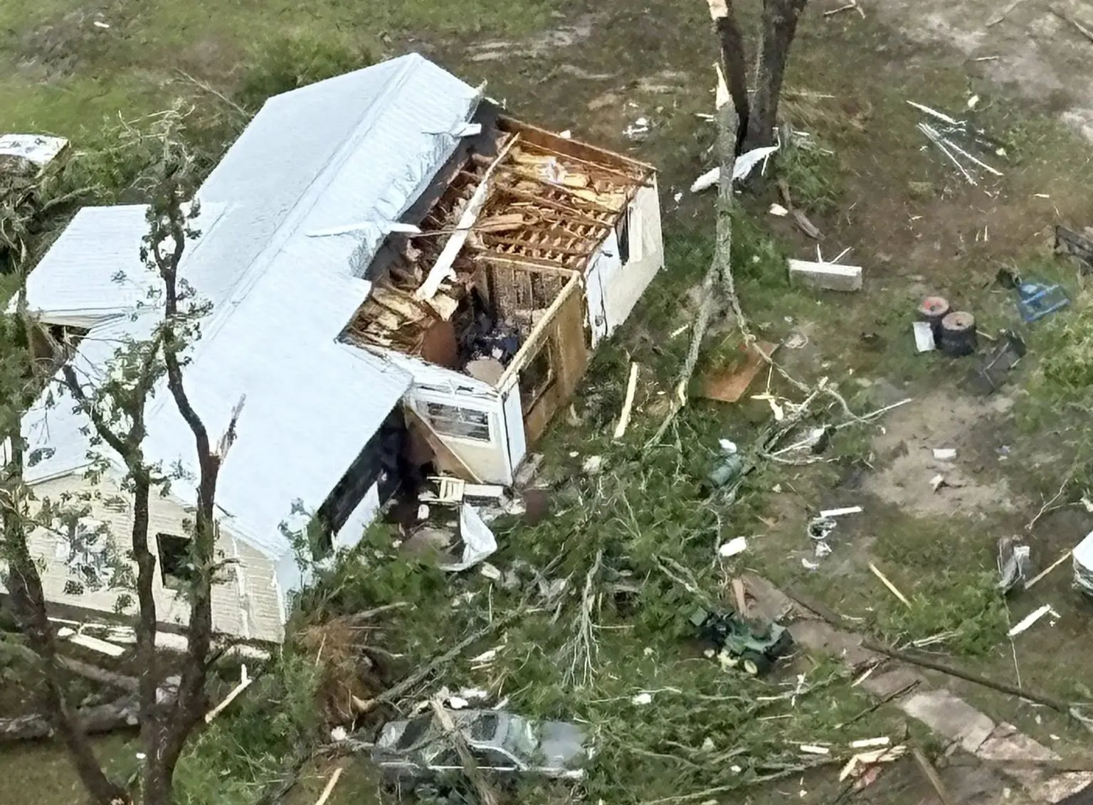 An aerial view shows a severely damaged mobile home surrounded by debris, fallen trees, and scattered objects after a tornado. A white SUV is parked nearby on a road littered with broken branches and other debris.