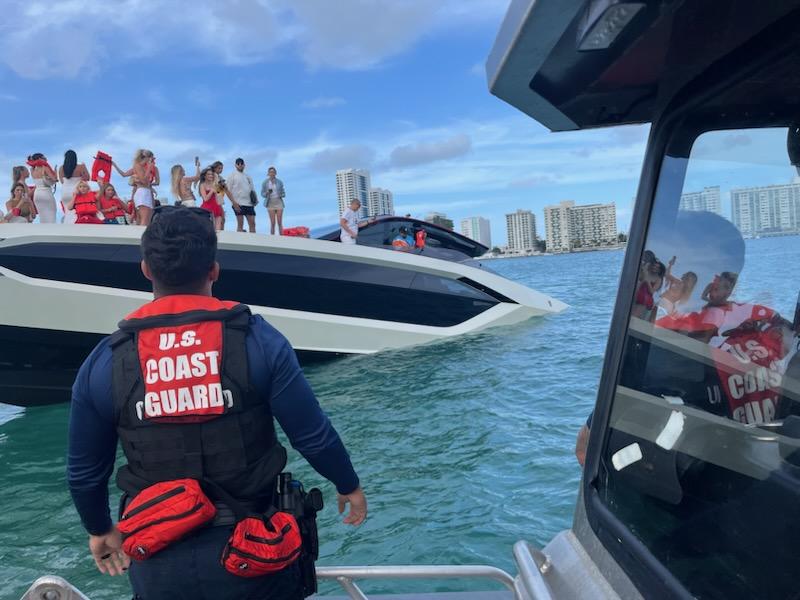 A U.S. Coast Guard member in a life vest stands on a vessel, observing a large yacht tilted heavily to one side in the water near Monument Island. Several passengers on the yacht, wearing life jackets, are visible on the deck, with high-rise buildings in the background under a partly cloudy sky. Another Coast Guard vessel is nearby, assisting in the operation.
