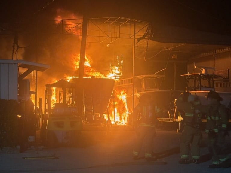 A nighttime scene at a boat yard in Tavernier, Florida, shows a large fire engulfing several boats. Bright orange and yellow flames illuminate the area, casting light on nearby structures and equipment. Two firefighters in full gear stand in the foreground, observing the blaze, while a yellow forklift is visible to the left. The sky is dark, and smoke rises into the air, partially obscuring the scaffolding and tarps overhead.