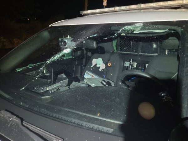 A nighttime photo of a Monroe County Sheriff's Office vehicle with a shattered windshield, illuminated by artificial light. Papers and equipment are visible inside the vehicle, and a person stands in the background near trees.