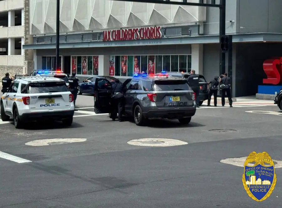 A street scene in downtown Jacksonville shows two police vehicles with flashing lights parked near a building with a sign that reads "Jax Children’s Chorus." Several officers are present, some near a gray SUV, while others stand nearby. A traffic light and street sign for Forsyth Street are visible above. The Jacksonville Sheriff’s Office logo is in the bottom right corner.