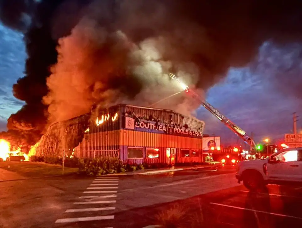 A large fire engulfs Southeast Pools & Spas in Port St. Lucie, with thick black smoke and orange flames rising into the evening sky. Firefighters use an aerial ladder to spray water on the burning building, while emergency vehicles and personnel are visible in the foreground. Credit: St. Lucie County Fire District