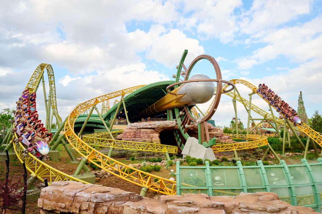 A dynamic aerial photograph of the Stardust Racers dual-launch roller coaster at Universal Epic Universe, capturing two vibrant trains—one purple with riders in red attire and another white—racing side-by-side along twisting yellow tracks elevated on green metallic supports. The cosmic-themed structure features a large golden spherical element and intertwining hoops amid rocky red terrain, lush green foliage, and a partly cloudy blue sky, with distant themed elements like a tall spire visible in the background.